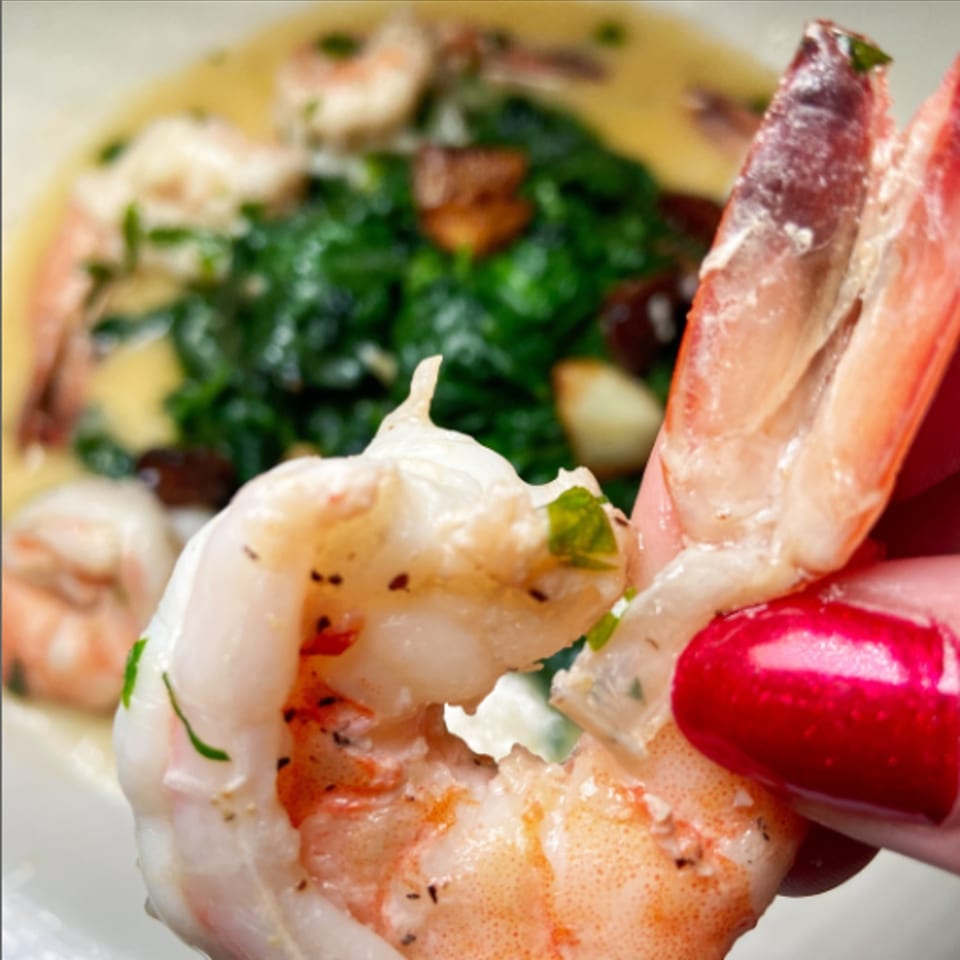 A hand with red nail polish holds a large shrimp over a dish of shrimp, spinach, and sauce.