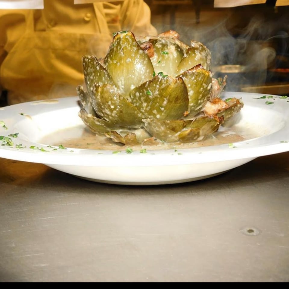 Steaming, whole baked artichoke in a white bowl with sauce, garnished with parsley.